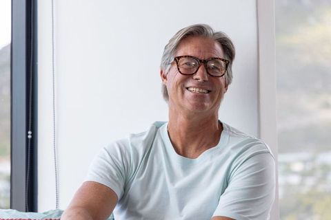 Smiling mature man wearing tortoiseshell glasses sitting by window in bright home interior