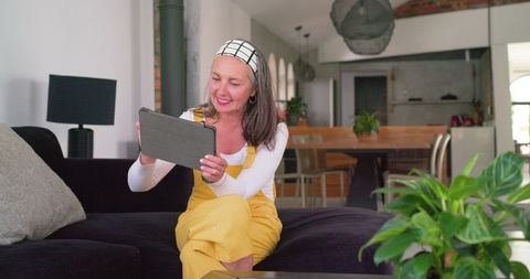 Smiling middle-aged woman using tablet sitting on sofa in bright open-plan living room