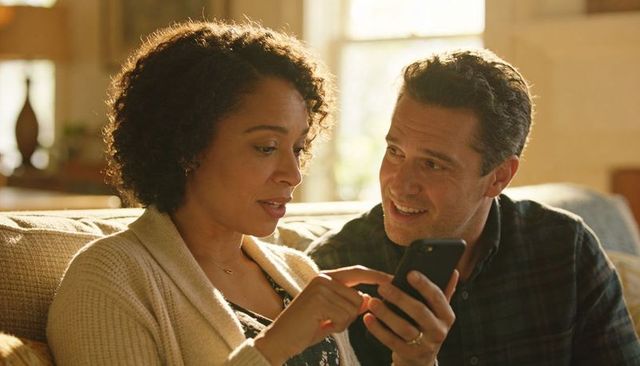 Midlife couple sharing smartphone moment on sunlit couch, tapping smiling in cozy home