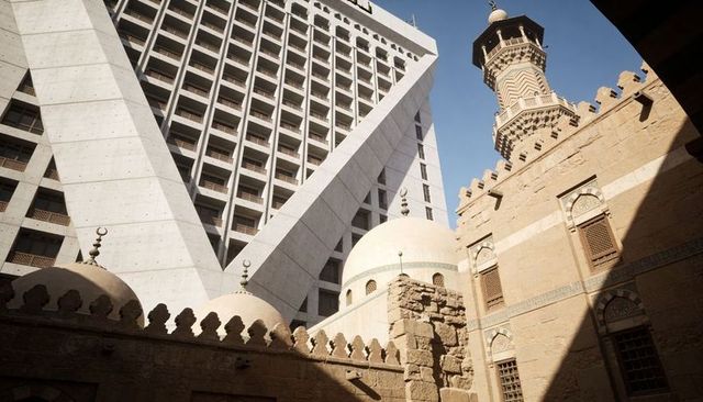 Triangular concrete tower contrasting with mosque domes and minaret in sunlit courtyard
