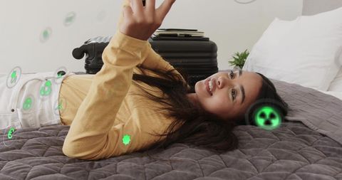 Young woman lying on bed using smartphone with augmented reality travel icons and suitcase