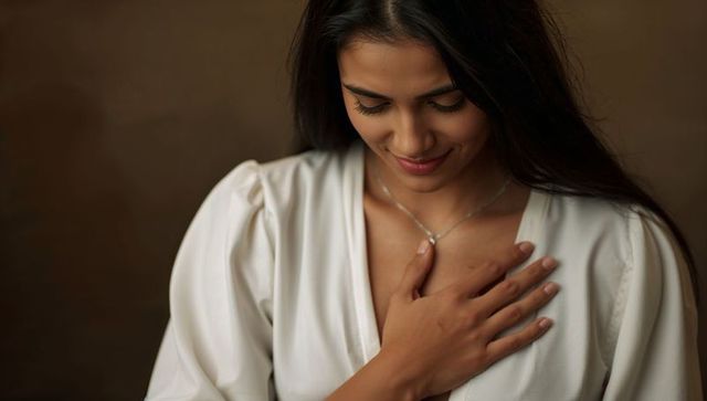 Smiling South Asian woman placing hand on chest wearing white blouse and delicate pendant