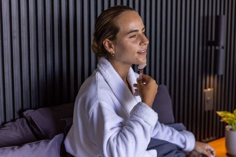 Woman in white bathrobe using rose quartz facial roller on jawline in minimalist bedroom