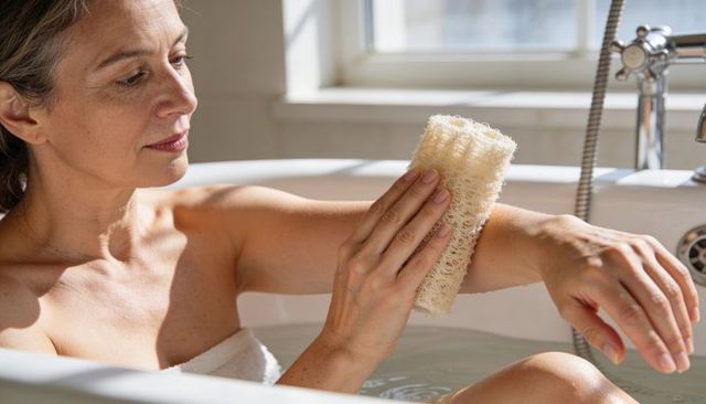 Mature woman enjoying relaxing bath with loofah in bright minimalist bathroom