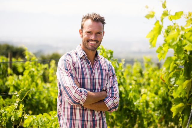 Man standing in vineyard with arms crossed, smiling confidently. Ideal for use in agricultural promotions, wine industry marketing, rural lifestyle blogs, and outdoor activity advertisements.
