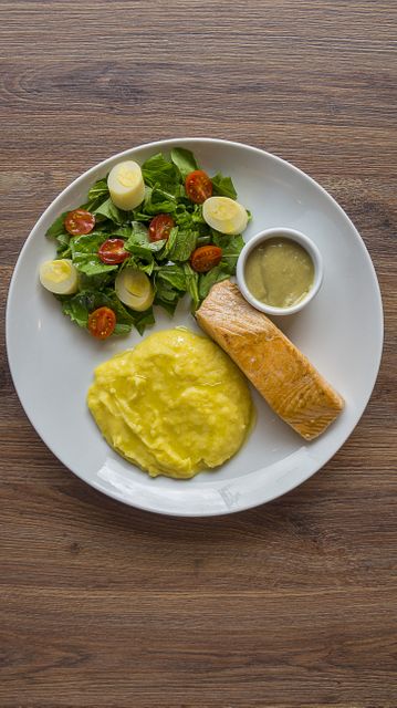 Healthy salmon meal on white plate featuring mashed potatoes, cherry tomato salad, and sauce. Suitable for websites or articles on healthy eating, nutritious recipes, gourmet food, or meal planning.