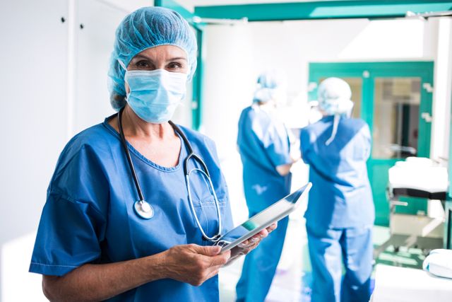 Surgeon in blue scrubs and mask using a digital tablet in a hospital corridor, with colleagues in the background. Ideal for use in healthcare, medical technology, and teamwork-related content.