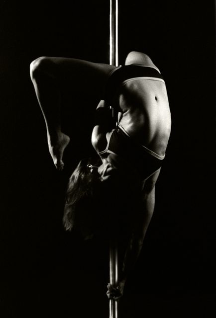 Woman demonstrating impressive acrobatic move on a pole in low light setting. Shows strength, flexibility, and artistry. Suitable for use in articles or promotions related to fitness, dance, gymnastics, performance arts, and women's empowerment.
