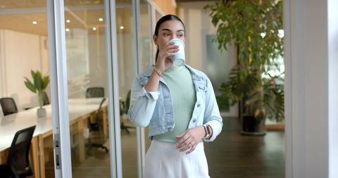Young professional woman drinking coffee in modern coworking office by glass meeting room