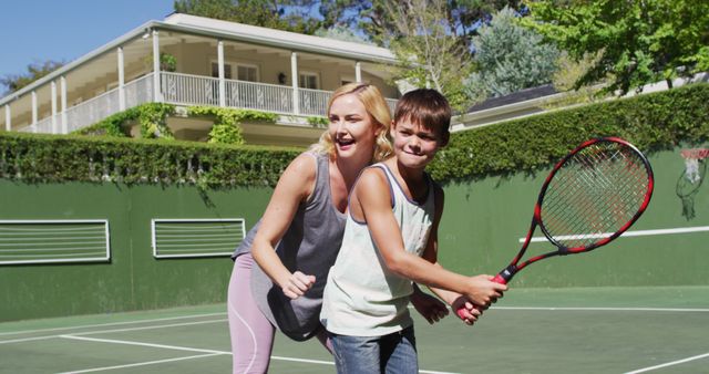 Mother and son spending quality time together learning tennis on an outdoor court on a sunny day. The scene is ideal for highlighting family bonding, active lifestyles, or sports training. Useful for outdoor or summer activity promotions and health and fitness materials.