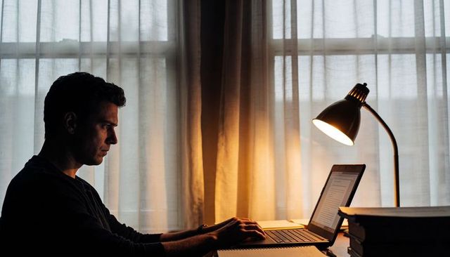 Late night focus: man typing on laptop under desk lamp in quiet home workspace