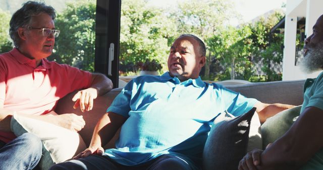 Three mature men sit on couch, engaged in relaxed conversation in sunlit living room. Suitable for lifestyle, senior community, friendship, and leisure concepts in advertising and editorial content.