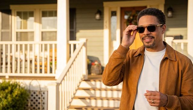 Adjusting sunglasses on sunlit porch steps wearing brown jacket and casual tee