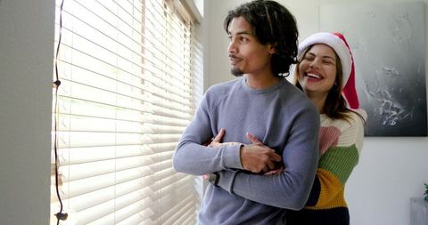 Couple Hugging by Sunlit Window Wearing Santa Hat Laughing in Cozy Living Room