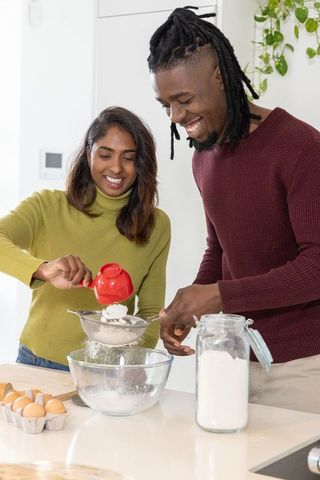 Diverse couple baking together in bright modern kitchen sifting flour and smiling