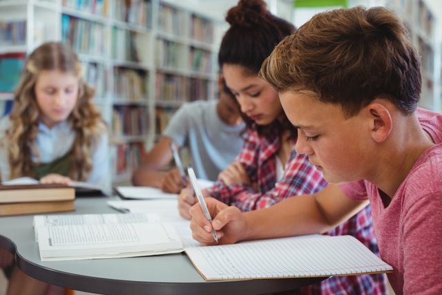 Students are engaged in studying at a school library, writing in notebooks and focusing on their work. The image is useful for educational materials, school brochures, learning resources, and articles on academic success and collaboration.