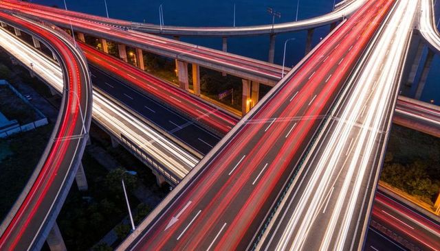 Nighttime elevated highway interchange showing dynamic red and white light trails