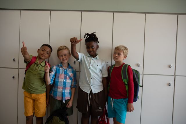 Multiracial elementary schoolboys standing in front of lockers in a school corridor, showing thumbs up. They are wearing casual clothing and backpacks, indicating they are students. This image can be used for educational materials, school promotions, diversity and inclusion campaigns, and childhood development articles.