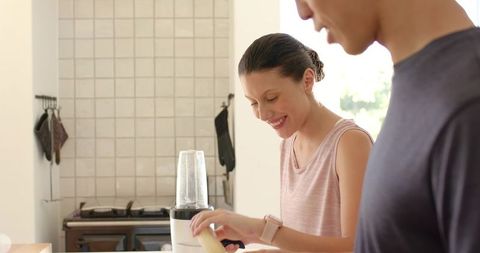 Couple preparing banana smoothie in sunlit kitchen smiling woman adding banana to blender