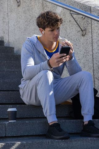 Teenage male sitting on concrete steps eating sandwich and scrolling smartphone