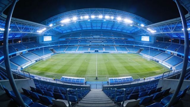 Floodlit modern stadium interior showing empty soccer pitch, goalposts and team benches