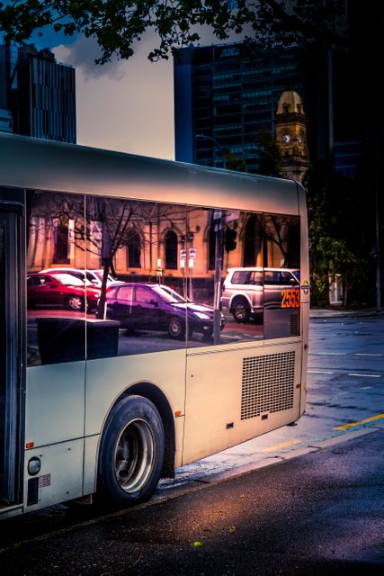 Image captures a city bus during evening with its windows reflecting street lights and nearby buildings. Picture showcases urban transportation and city life, illustrating the hustle and bustle of downtown streets. Ideal for articles or blogs about urban commutes, public transportation, or city planning.