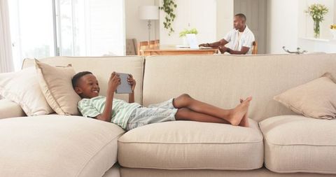 Child relaxing on beige sofa using tablet while parent working remotely at dining table