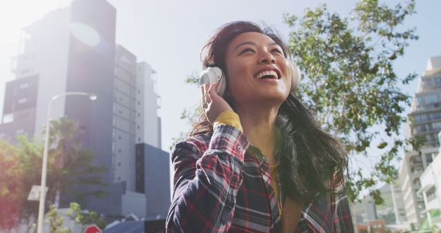 Smiling asian woman wearing headphones listening to music in sunny city street. City living, relaxation, music and modern urban lifestyle.
