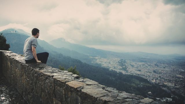 Man sitting on stone wall contemplating a scenic mountainous landscape overlooking a city. Cloudy sky enhances the serene and tranquil atmosphere, perfect for usage in topics related to travel, solitude, meditation, nature, mental wellbeing, wanderlust, and tourism promotions.