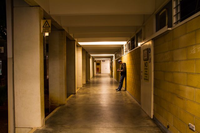 Man leaning against a wall in a quiet, empty hallway at night. He appears deep in thought, standing in a dimly lit corridor. Indoor scene with an industrial setting featuring concrete floors and yellow walls. Useful for depicting solitude, contemplation, loneliness, urban lifestyle, night environment themes, and architectural spaces.