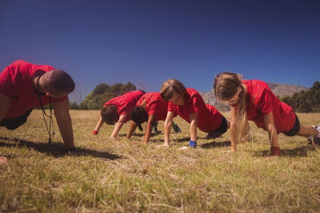 Trainer leading group of kids in outdoor boot camp exercise on a sunny day. Children performing push-ups on grass, emphasizing teamwork, physical fitness, and outdoor activity. Ideal for use in articles about children's fitness, summer camps, outdoor activities, and team-building exercises.