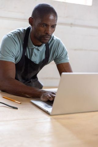 Mature african american craftsman leaning over workbench typing on laptop