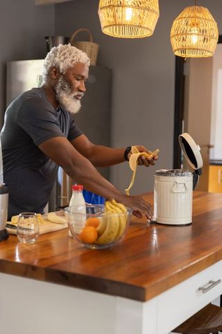 Senior african american man placing banana peel into white pedal bin on kitchen island