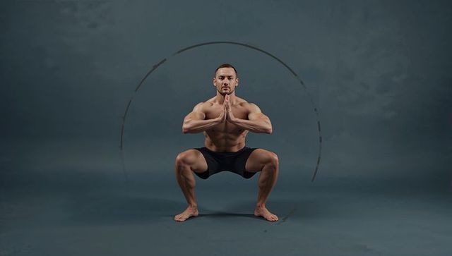 Muscular male holding deep squat prayer pose training barefoot in studio with arc graphic