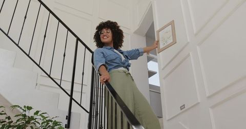 African american woman hanging framed art on white paneled wall next to staircase