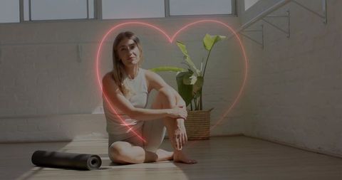 Sitting woman hugging knee on sunlit loft floor with neon heart and yoga mat