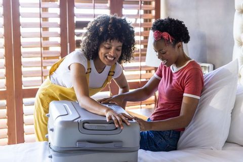Black women friends packing gray hard-shell suitcase on sunlit hotel bed, smiling together