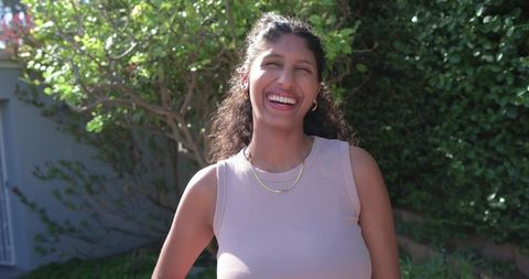 Laughing woman standing in sunny backyard wearing tank top, gold necklaces, hoop earrings