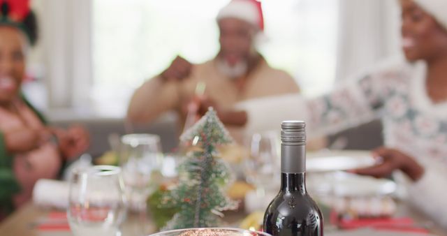 Family enjoying Christmas festivities around a dinner table. The focus is on a bottle of wine, with blurred family members wearing Santa hats in the background. Perfect for holiday greeting card designs, festive campaign promotions, and seasonal social media posts.