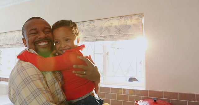 African American father hugging his young daughter in a bright, sunlit kitchen. Both are smiling, radiating happiness and affection. Sunlight filters through the window, adding a warm, glowing effect. This can be used to highlight themes of family love, parental bonding, joy, and the warmth of everyday moments.