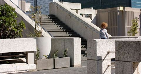 Sunlit urban woman standing on concrete plaza steps carrying crossbody bag