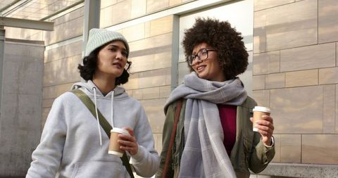 Two friends walking and holding takeaway coffee on urban sidewalk in casual winter outfits