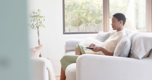 An African American woman is sitting comfortably on a sofa, using a tablet in her hands, with modern home decor in the background and natural light coming through the window. This can be used for content related to home lifestyle, modern technology use at home, relaxation and leisure activities, and home interior designs.