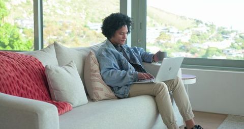 African american man working remotely on laptop in cozy living room with window view