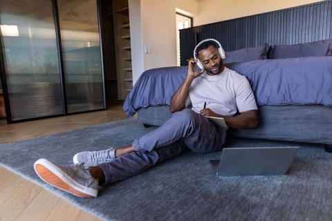 African american man working remotely from bedroom wearing headphones and writing notes
