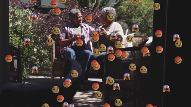 Senior couple sits on terrace holding coffee mugs, surrounded by varied emojis, conveying emotions. Bright, sunny outdoor setting emphasizes relaxation. Ideal for projects focusing on digital communications, crypto cultural humor, or highlighting aging lifestyle enjoyment intersecting with modern virtual trends.