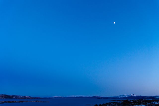 Serene twilight view with a bright moon hanging in a clear blue sky over a calm ocean. Distant mountains and coastline frame the horizon. Ideal for backgrounds, travel promotions, serene landscape themes, and inspirational nature content.