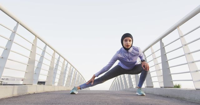 Fit biracial woman wearing hijab and sports clothes stretching legs on city bridge. City living, fitness and healthy modern urban lifestyle.