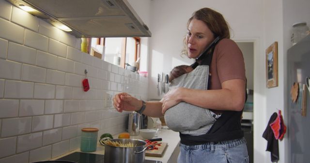Image of caucasian mother with newborn baby in baby carrier using smartphone in kitchen. motherhood, parental love and taking care of newborn baby concept digitally generated image.