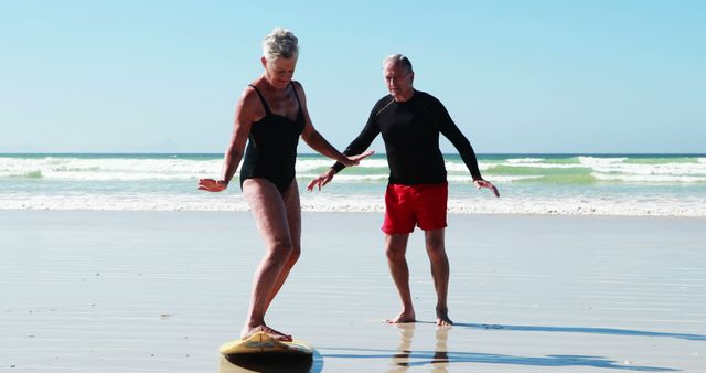 A senior Caucasian woman balances on a surfboard on the beach while a senior Caucasian man assists her, with copy space. Their activity suggests they are enjoying a leisurely day at the beach, learning to surf together.